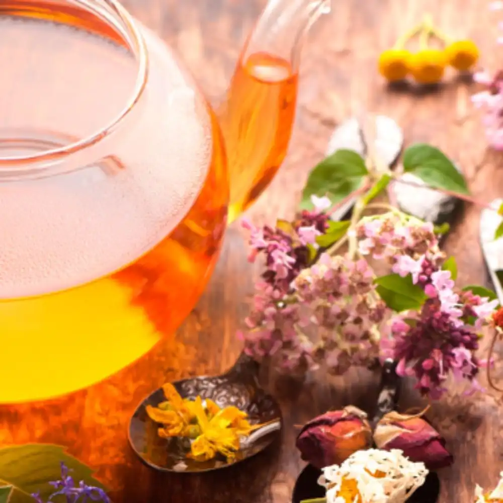 A clear glass teapot with amber herbal tea, surrounded by dried lavender, chamomile flowers, and botanical ingredients on a wooden surface.
